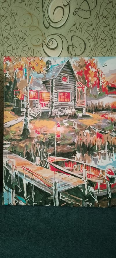 This is a painting of a rustic log cabin situated near a body of water, likely a lake or river, in the village of Абалак, Russia. The scene depicts an autumn setting, with trees displaying vibrant red and yellow foliage in the background, suggesting a cool, crisp day. The cabin itself is made of dark logs with red-accented windows and a porch. In the foreground, a wooden dock extends into the water, where two red boats are moored. The water reflects the surrounding landscape and the boats, adding depth to the composition. A lantern with red shades is positioned on the dock, casting a warm glow. The overall mood is tranquil and serene, evoking a sense of peaceful country living. There is no visible text in the painting.