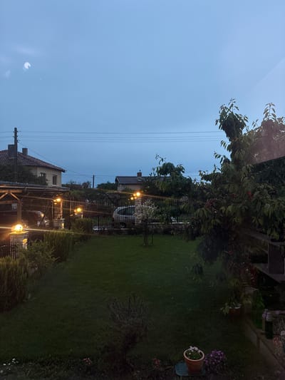 The image captures a serene evening view of a residential garden and neighboring houses, likely taken from an elevated perspective such as a window. The scene is illuminated by the fading light of dusk or early evening, characterized by a clear, light blue sky that gradually darkens towards the horizon. A prominent, crescent-shaped reflection or glare is visible in the upper left sky, suggesting the photograph was taken through glass.
In the foreground, a well-maintained green lawn spans much of the lower frame. Lush greenery dominates the right side, featuring a large tree with dark green leaves and clusters of small red berries. Various shrubs and bushes are dispersed throughout the garden, and a terracotta pot with white flowers, alongside another with purple blooms, adds color to the far right.
The mid-ground reveals several houses with traditional red-tiled roofs. A black metal fence or gate separates the immediate garden from the adjacent properties. Numerous outdoor lights, including lanterns and string lights, cast a warm, yellow glow, particularly around a pergola-like structure and along paths, creating a cozy ambiance. A white vehicle is partially visible behind the fence near one of the houses. Utility poles and power lines are noticeable in the background and mid-ground against the twilight sky.
The scene is devoid of human activity, conveying a sense of peace and tranquility. There is no visible text. The architectural style and garden elements are consistent with the provided location context of General Kantardjievo, Bulgaria.