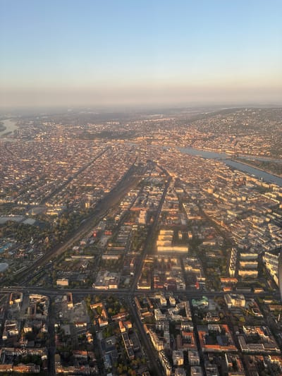 The image shows an aerial view of Budapest, Hungary, during daytime with clear skies and visible atmospheric haze in the distance. The cityscape features dense urban development, consisting of buildings of varying heights and styles. A river runs through the city, with multiple bridges spanning its width. Train tracks and roads are visible, indicating transportation infrastructure. The lighting suggests a late afternoon or early morning, casting shadows on the buildings. The exact location within Budapest cannot be determined without further information. Vecsés is a separate city outside of Budapest, and there is no evidence in the image that it is Vecsés.
