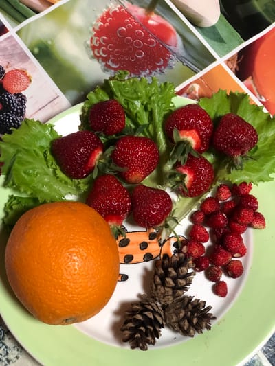 A close-up, top-down shot displays a light green ceramic plate holding a selection of fruits and pine cones. The plate is filled with fresh strawberries, some whole and some halved, arranged on a bed of bright green lettuce leaves. To the left of the strawberries, a whole orange sits prominently. A cluster of tiny wild strawberries is scattered to the right of the larger strawberries. Two brown pine cones rest on the lower portion of the plate, next to a decorative cartoon drawing of a ladybug. The background features a collage of fruit-themed images, including strawberries and other berries. The scene is brightly lit, highlighting the vibrant colors of the food. The image does not contain any people or provide any information about the location in Staroe Istomino, Russia.