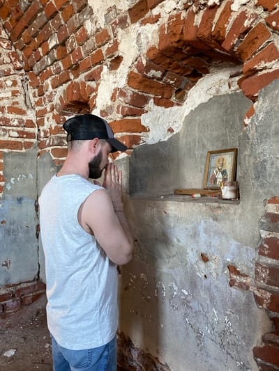 A man wearing a grey trucker hat, a white t-shirt, and blue jeans is shown praying in a small niche or alcove, likely within a building or structure. The walls of the alcove are constructed of red bricks, some of which are damaged or deteriorating. A framed religious icon or painting, potentially of Christian saints or religious figures, is mounted on the wall above the man's head. A small shelf or wooden support structure is placed beneath the icon. The location is potentially in Шуран, Russia.