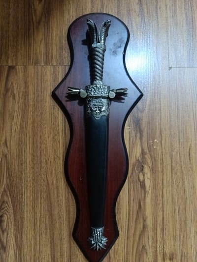 A decorative dagger with a black sheath and ornate silver handle is mounted on a dark wooden plaque. The plaque is shaped like a stylized shield and is attached to a wood-paneled wall. The dagger's handle features intricate carvings of serpents and a face. The background shows a wooden wall with vertical planks. The image does not contain information about the city of Yukariyuz, Uzbekistan.