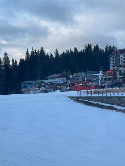 The image presents a wide view of a ski resort base area in Stoikite, Bulgaria, under overcast, dim lighting conditions suggestive of dusk or early evening. In the foreground, a vast expanse of groomed snow defines a ski slope. A low stone wall with a dark metal railing extends across the mid-right, separating the lower snowfield from an elevated section.

On this elevated section, a cluster of resort buildings is visible, including multi-story structures with various architectural styles, some featuring balconies. Prominent signage, including "HEAD" flags and a partially visible red neon sign (possibly "HAPPY END"), adorns these establishments, which appear to be restaurants, bars, or shops. Outdoor seating areas with wooden tables and benches are arranged in front of several buildings.

A large, red and white tracked snow groomer is positioned on the snow near the base of the buildings, facing right. Several indistinct human figures are visible near the buildings and outdoor areas. The background is dominated by a dense forest of dark evergreen trees covering a steep mountainside behind the resort structures. The sky is largely grey and overcast, with blurred light reflections, suggesting the photograph was taken through glass.