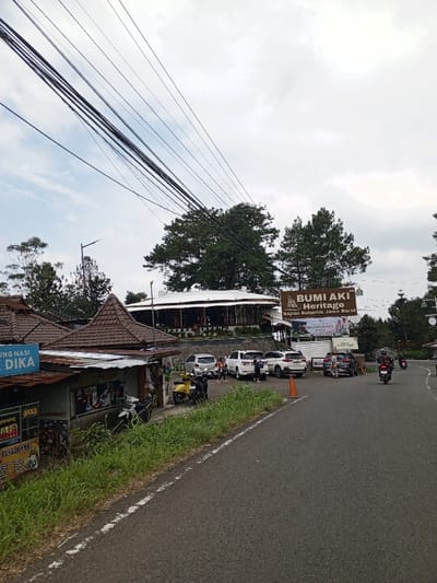 The image depicts a roadside scene in Ciloto, Indonesia, under an overcast sky. Numerous dark electrical and communication wires span across the upper left portion of the frame. An asphalt road, with a white dashed line on its left edge, curves slightly to the right, receding into the background.
On the left side of the road, a grassy verge separates the road from a series of small, rustic single-story buildings. These structures feature peaked brown tiled roofs and corrugated metal awnings. One building displays a partially visible sign that reads "RUNG NASI BU DIKA." Several motorcycles are parked near these buildings, including a yellow scooter and two dark-colored scooters.
To the right of the road, an orange traffic cone is positioned near an elevated parking area. This parking lot contains multiple parked vehicles, including several white SUVs, a silver sedan, and other dark-colored vehicles. Several individuals, consisting of both adults and children, are observed standing and moving within this parking area, some near the parked cars and others closer to an entrance.
Behind the parking lot, a large, prominent brown sign with white text identifies "BUMI AKI HERITAGE" and "SAJIAN AUTENTIK JAWA BARAT." Further back and higher in elevation, the main structure of Bumi Aki Heritage is visible: a large, circular building with a distinctive white roof and dark-paneled walls, surrounded by dense green evergreen trees.
In the mid-distance on the road, two motorcycles are present, with one actively moving away from the viewer. The background features additional evergreen trees and distant hills partially obscured by atmospheric haze. A street light pole is visible on the left side, behind the smaller buildings.