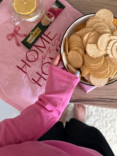 This media file shows a top-down view of a person's arm and hand reaching for a bowl of potato chips. The setting appears to be indoors, possibly a home, given the presence of a wooden table, a white fluffy rug, and a pink textured cloth with "HOME" written in a decorative script. The person is wearing a bright pink long-sleeved shirt.
In the foreground, there's a glass mug with a slice of lemon and some clear liquid, likely water or tea. Next to it are two small packages, one with the text "NEW" and an image of berries and cream, and another green package with the text "MONARCH" and a coffee bean logo. The bowl is filled with many round, thin potato chips.
The person's hand, with light blue dotted nail polish, is reaching into the bowl. The overall mood seems relaxed and casual, suggesting a moment of snacking or taking a break. The lighting is bright, implying daytime. There are no obvious cues about the weather. The location context provided is Tliustenkhabl', Russia.