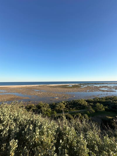 The image shows a beach scene in Vila Real de Santo António, Portugal. The sky is clear and blue. To the right of the sky is the shore with water and green vegetation and trees.