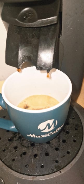 A dark-colored capsule coffee machine is dispensing a hot beverage into a teal mug on a black, perforated drip tray, likely within a domestic or commercial indoor setting in Vendin-le-Vieil, France. Two streams of light brown liquid with visible steam or vapor are actively flowing from the machine's twin spouts into the mug. The mug, which has a white interior, contains a small volume of coffee with a light brown crema layer and some darker residue. A white logo, consisting of a stylized "M" above the word "MaxiCoffee," is prominently displayed on the side of the mug. The drip tray exhibits dark liquid splashes and residue. The upper left side of the coffee machine features a circular depression with an embossed design.