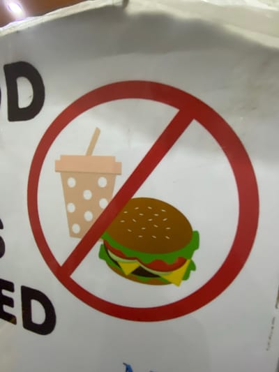 The image shows a "no food or drinks" sign at the St. Joseph the Worker Parish Church in San Jose del Monte, Philippines. The sign is a red circle with a diagonal line through it, indicating prohibition. Inside the circle are two images: a cartoon of a beverage cup with a straw and a cartoon of a hamburger. The sign is mounted on a white surface, possibly a wall or a bulletin board, with some text visible above and below the sign. The text above appears to be part of the word "FOOD," and the text below includes the word "ED." The lighting suggests an indoor setting, possibly with overhead lights casting reflections on the surface. The overall impression is a clear directive to refrain from consuming food and beverages within the church premises.