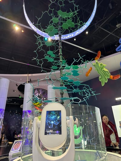 This image depicts an indoor exhibition hall in Guangming District, China. Central to the scene is a large, multi-layered sculpture composed of green molecular models, suspended from a dark ceiling. Above this sculpture, a curved, translucent ring with internal blue illumination displays the fragmented text "TERIOUS MEDICAL HELICAL STRUCTU." Directly below the suspended structure, a white, vertically oriented interactive touchscreen kiosk displays a vibrant molecular illustration and Chinese text on its screen, set upon a clear cylindrical base. To the right, various oversized, colorful models of cellular components, including an orange, elongated structure and a green, convoluted one, are suspended from above. A female individual, with dark hair, wearing a dark red jacket over a white top, is partially visible on the far right, positioned near a clear display, her gaze directed downwards. On the left, tall, white cylindrical columns emit a soft purple glow, adjacent to a spherical, illuminated exhibit further in the background. The ceiling is dark, revealing exposed grids and recessed lighting fixtures.