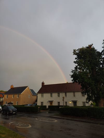 A rainbow arcs across a gray, overcast sky over a residential street in the town of Needham Market, Mid Suffolk, United Kingdom. The street, which appears wet and reflective, has painted speed limit signs on the asphalt. On the left side of the street, a light-colored house with a dark roof and multiple windows is visible, with a dark-colored car parked in front. To the right of this house, a larger, white-sided house with a reddish-brown roof and numerous windows stands, with a tall, leafy tree to its right. Hedges line the front of the houses. The sky is muted, suggesting a recent or impending rain shower.
