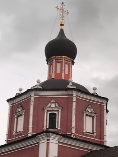 A red domed church stands against a cloudy sky. The church has a black dome, topped with a golden cross. It features ornate white trim and windows. The church is located in the city of Ozeretskoe, Russia.