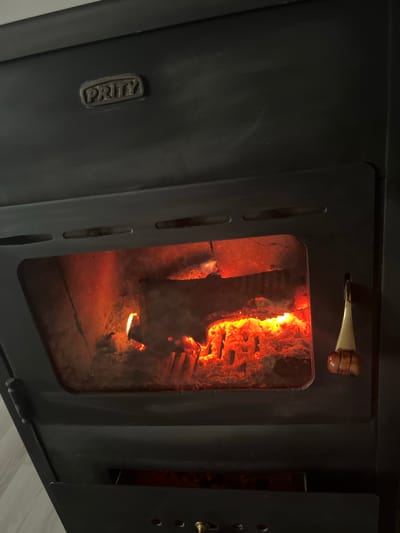 The image shows a black wood-burning stove with a fire burning inside. The stove has a glass door, partially obscured by the flames and embers. The handle for opening the door is visible at the top. The brand "PRITY" is embossed on the left side of the stove. The setting seems to be indoors. There is no indication of the location being Sredkovets, Bulgaria, or any details about people or actions.