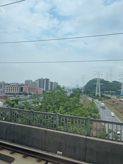 The image shows a view from a train in Dongguan, China, with a cityscape in the distance and a highway running parallel to the tracks. The cityscape features several tall apartment buildings and other structures, suggesting a densely populated urban area. A multi-lane highway is visible, with cars and trucks traveling along it. Large electricity pylons are scattered throughout the landscape, indicating the presence of significant power infrastructure. Lush green trees and vegetation line the highway and surround the pylons, contrasting with the built environment. The sky is overcast with clouds, and power lines are visible crossing the top of the frame. The foreground of the image shows the train tracks and a concrete barrier with a decorative metal fence. The scene suggests a mix of urban development, transportation networks, and natural elements, typical of a modern Chinese city.