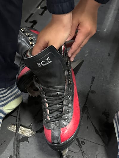 A pair of hands is actively engaged with a black and red high-top boot or skate. The person's forearms, covered by dark sleeves, are visible as they manipulate the footwear. The boot itself is predominantly black with a distinct bright red section across the toes and sides. It features black laces threaded through metal eyelets and hooks. White letters spelling "S.C.E" are visible on the black cuff of the boot.
Protruding from the sole of the boot is a blade-like attachment, indicating it is likely an ice skate or specialized boot. This blade appears somewhat worn or corroded. A striped sock, light green/yellow and dark blue/black, is partially visible near the bottom right, suggesting the person is either wearing or about to wear the skate.
The activity appears to involve adjusting the boot, possibly lacing it up or preparing it for use. The setting is a dark, possibly industrial or utilitarian area, with the floor showing streaks and droplets of water or other liquid, which is consistent with environments where ice skates are used or maintained. The image was captured in Garges-lès-Gonesse, France.