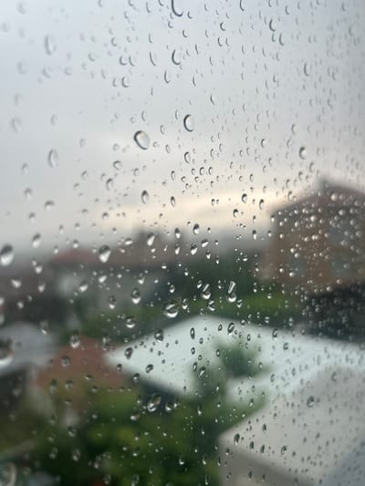 The image displays numerous raindrops of varying sizes adhering to a transparent surface, most likely a window or windshield. The water droplets in the foreground are in sharp focus, showcasing their individual shapes and reflections of the ambient light.

Behind the rain-covered surface, the background is significantly blurred, indicating a shallow depth of field. Indistinct shapes on the left and center suggest structures or buildings with muted grey and brownish tones. On the right side, blurred patches of green foliage, likely trees or bushes, are visible. The overall lighting suggests an overcast sky during daytime.

The scene clearly depicts rainy weather conditions. The perspective is from an interior looking outwards, creating a contemplative mood through the obscured view. There is no visible text in the image. This scene is set in Aytos, Bulgaria.