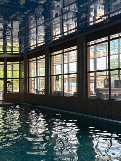 This image depicts an indoor swimming pool facility located in Tulovo, Bulgaria. The foreground is dominated by a rectangular swimming pool filled with light blue-green water, its surface actively rippling and reflecting the overhead structures and outdoor light. The pool's perimeter features a dark tiled edge, with a lighter blue strip directly below it, and a dark, possibly inflatable object resting on the edge in the middle-right. A metallic handrail is present on the left side of the pool.
A wall constructed of small, variegated brown and beige square tiles forms the background to the pool, extending upwards to a series of large, dark-framed windows. These windows illuminate the space with natural light and reveal an outdoor setting. Through the windows, lush green trees and foliage are visible, particularly on the left and right. Outdoor seating areas, comprising tables and chairs, are discernible, with faint human figures appearing to be seated at some tables in the mid-ground. A potted plant with green leaves stands to the left of the leftmost window. To the right, an outdoor counter or bar area is visible, featuring a monitor displaying a human face, and dark furniture or storage boxes beneath it. A red-roofed structure and a blue canopy are also visible through different window panes. A circular object, possibly a wall clock, is reflected on the ceiling and also visible through the windows on an interior wall beyond the outdoor area.
The ceiling is highly reflective, appearing dark blue with lighter, abstract patterns resembling clouds, and acts as a mirror, reflecting the window grid, the outdoor environment, and various indoor elements in an inverted and distorted fashion. The indoor area receives significant natural illumination from the exterior, creating a contrast with the relatively shadowed interior.