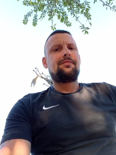 A selfie photograph shows a man from a low angle, taken outdoors. The man has short, dark hair, a beard, and a piercing above his right eye. He is wearing a black t-shirt with a white Nike swoosh logo on the left chest. Behind him, leafy branches and a light blue sky are visible. The specific location in Lormont, France cannot be determined from the image alone.