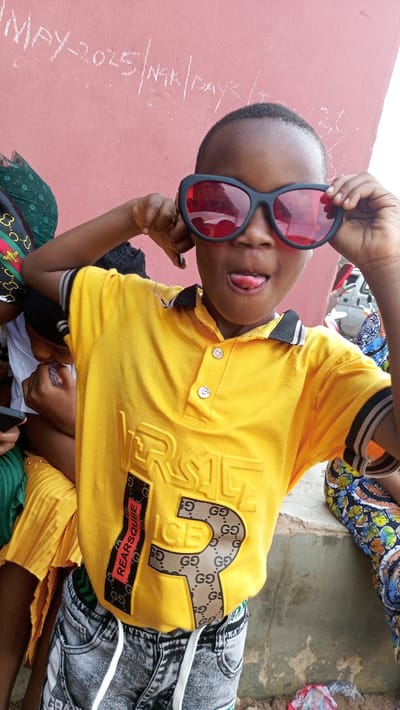 A young child wearing a yellow polo shirt, featuring "Versace" text and a Gucci-patterned number, is posing with red-tinted sunglasses. The child is sticking their tongue out, adding to the playful expression. They are wearing gray shorts and are surrounded by other individuals. A wall with handwritten text is visible in the background.
