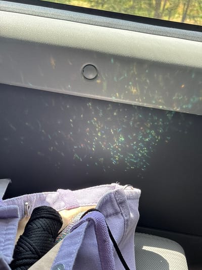 The image displays a close-up view of a car's interior, focusing on a light-colored, textured dashboard and a portion of a purple canvas tote bag. Scattered across the dashboard are numerous small, shimmering reflections of light, appearing as colorful speckles of green, blue, yellow, and red. A circular indent, possibly a button or a small compartment, is visible on the dashboard. The tote bag, partially visible in the foreground, contains a tightly wound spool of black cord. The background, visible through the car window, shows blurred greenery, suggesting an outdoor environment. The overall scene captures the effect of sunlight interacting with the car's interior surfaces. The location is not specified in the image.