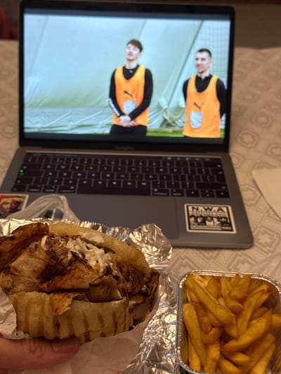 A close-up, slightly angled shot captures a person holding a large, partially wrapped kebab in their left hand. The kebab appears to be made with grilled meat, lettuce, and sauce, tucked into a pita or flatbread. To the right of the kebab, a small foil container is filled with golden-brown french fries. In the background, a laptop displays two young men in orange vests and black long-sleeved shirts. The laptop is placed on a textured, light-colored surface. There is no discernible connection to Ravenna, Italy.