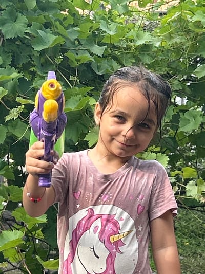 The image shows a young girl holding a purple and yellow toy water gun, looking towards the camera with a slight smile. Her hair is wet, and she is wearing a pink t-shirt with a unicorn design. The setting appears to be outdoors, with lush green foliage in the background, suggesting a natural or garden environment. The lighting indicates it might be daytime, possibly sunny. The girl's expression and the toy water gun suggest she is engaged in play, perhaps during a warm-weather activity. The image is framed such that the girl and the water gun are the main focus, with the greenery providing a soft, natural backdrop. The orientation of the image is rotated.