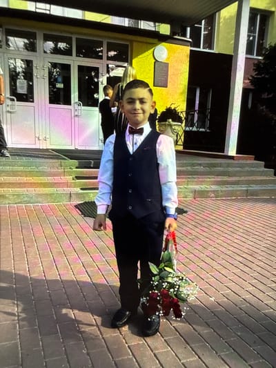 A young boy, dressed in a formal outfit with a white shirt, dark vest, and a maroon bow tie, stands on a paved pathway in front of a building. He is holding a bouquet of dark red roses and small white flowers. The building appears to be a school, with large glass doors and windows, painted in yellow and brown. Steps lead up to the entrance. The pathway is made of brick pavers, with shadows cast by sunlight. The overall scene suggests an event, possibly a first day of school or a ceremony, taking place outdoors in a city.