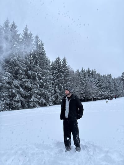 A male subject stands in a snow-covered landscape in Le Hohwald, France, under an overcast sky during active snowfall. The individual, appearing approximately 20-30 years old, has a dark beard and wears a grey knitted beanie, a black padded jacket over a light grey hooded sweatshirt, and black snow pants with dark footwear partially submerged in the snow. He stands with hands in his jacket pockets, facing slightly right of center while looking towards the viewer's left. The ground is covered in deep, white, undisturbed snow. Behind the subject, a dense line of snow-laden evergreen trees extends across the frame. In the distant right, several indistinct figures and dark objects are visible on a snow-covered slope, suggesting recreational activity. Numerous blurred white specks are present throughout the image, consistent with falling snowflakes.