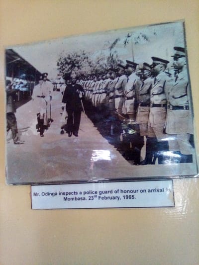 A black and white photograph, appearing to be an archival print under a reflective protective surface, depicts Mr. Odinga inspecting a police guard of honor on his arrival from Mombasa, 23rd February, 1965, at a location within the city of South Sakwa ward, Kenya. The scene shows Mr. Odinga, dressed in a dark suit and possibly holding a cane, walking from left to right along a line of approximately ten uniformed police officers standing at attention. The officers wear light-colored uniforms, dark peaked caps, and dark ties. An official in a light-colored uniform with a peaked cap walks slightly behind and to the left of Mr. Odinga. A partially visible individual stands on the far left near a covered platform or building structure. The event takes place outdoors on a paved or concrete surface, with trees visible in the background.