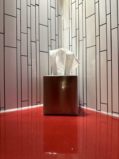 The photograph shows a tissue box dispenser affixed to a tiled wall next to a red partition. The tiles are white with dark grout, laid in a horizontal, staggered pattern. The tissue box is dark brown and contains white tissues. The red partition is smooth and glossy, with horizontal lines that suggest a similar tile pattern, though the lines are more faint. The setting is in the city of Hollywood Seminole Reservation, United States of America.