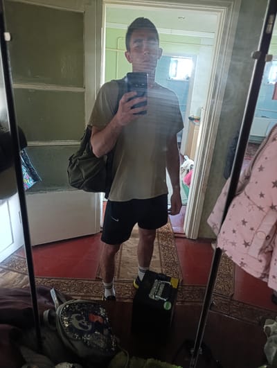 A young man is reflected in a mirror, taking a selfie. He is wearing a light brown t-shirt, black shorts, and white socks with yellow-soled shoes. He has a dark green backpack slung over his left shoulder and is holding a smartphone in his right hand, angled to capture the reflection.
The background visible in the mirror suggests an indoor setting. To the left, a door with glass panes is visible, with some items hanging on it. The floor is covered with a red rug with an intricate pattern. To the right, a pink jacket with star patterns is hanging, and further back, a washing machine and some other household items can be seen. The overall impression is of a room within a residential building. The image has been identified as being taken in Fastiv, Ukraine.