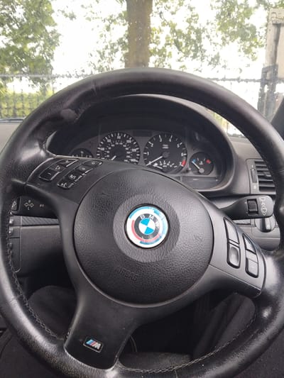 An interior shot of a car, focusing on the steering wheel and a portion of the dashboard, is captured from the driver's perspective. No people are visible in the frame.
The central subject is a black, leather-wrapped steering wheel, showing signs of wear on its surface. The iconic BMW logo, featuring its blue, white, and red segments, is prominent in the center of the airbag cover, which also has the word "AIRBAG" embossed. An "M" badge (associated with BMW M series) is affixed to the right spoke of the steering wheel. Integrated controls are present on both sides of the steering wheel's hub. The left cluster includes buttons for volume control (+ and -), an "R/T" button, and another with a speaker icon. The right cluster features buttons with arrow icons and a "1/0" button.
Behind the steering wheel, a section of the instrument cluster is visible, displaying a tachometer marked "x1000 r/min" and a speedometer with markings up to "240" (likely km/h, with smaller "MPH" markings visible up to "140"). Other gauges include a temperature gauge with red and blue zones, and a fuel gauge marked "1/2". A control stalk for wipers and washers is visible on the right side of the steering column. A small area of carbon fiber trim is also visible beneath the right steering wheel spoke.
Through the windshield, a bright, blurred view of green trees and light sky indicates it is daytime. The soft focus and potential droplets on the glass suggest either rain or condensation on the exterior of the vehicle, contributing to the ethereal background. The car appears to be stationary, possibly in Stafford, United Kingdom.