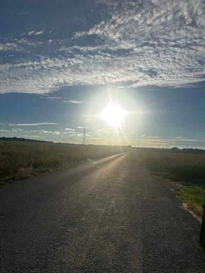 An eye-level, medium shot captures a desolate, paved road stretching into the distance under a bright, sun-drenched sky in Borough of Swale, United Kingdom. The sun, a radiant star with prominent rays, dominates the upper center of the frame, casting a warm glow across the scene. A line of white, wispy clouds stretches horizontally across the blue expanse, creating a textured canopy. The road, appearing to be a country lane, is bordered by fields of tall, dry grass that reach to the horizon. In the middle distance, a solitary, tall electricity pylon stands sentinel, its metallic structure stark against the natural landscape. Further back, a faint outline of trees and more pylons suggests a rural, possibly agricultural, setting. The overall impression is one of solitude and open space, with the sunlight accentuating the textures of the road and the surrounding vegetation.