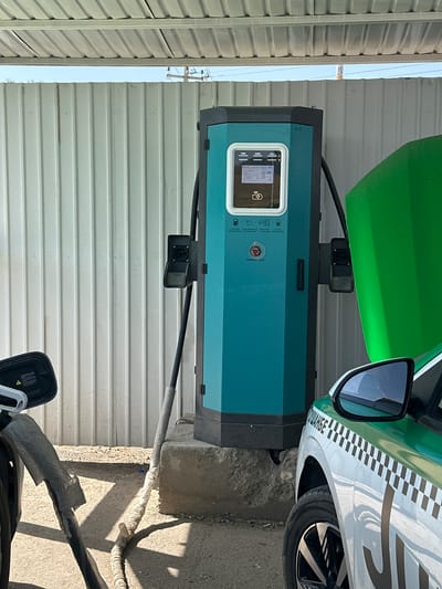 The image shows an outdoor electric vehicle charging station in Розиён, Tajikistan. A teal-colored charging station with a digital display is positioned centrally. Two charging cables are visible, one leading from the station towards the left side of the frame and another, partially obscured, to the right. A green car with a checkered pattern on its side is parked to the right of the charging station, with its hood open. The background consists of a corrugated metal wall and a portion of a metal roof. The ground is a mixture of asphalt and gravel. The scene appears to be a public charging point.
