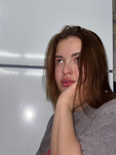 A young woman with light brown hair and fair skin is shown. She has a nose ring and is wearing a gray t-shirt. Her left arm is bent, and her hand is supporting her face. The background is a white surface with a vertical metallic strip. The location is unspecified.