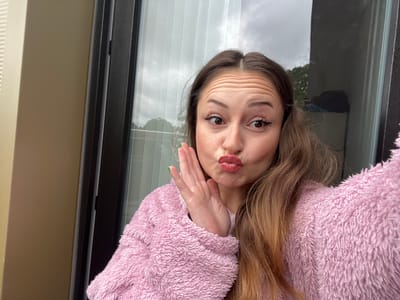 A young woman is featured in a close-up, chest-up selfie, captured in what appears to be Heusden-Zolder, Belgium. She is making a playful expression with her lips pursed into a "kiss face" and her eyes wide, looking slightly to her right. Her eyebrows are raised, creating visible wrinkles across her forehead. Her right hand is raised to the side of her face, with green nail polish visible on her fingernails.

She has long, light brown hair styled with a central part, and she is wearing a fluffy, light pink top or jacket. Her makeup includes defined eyebrows, winged eyeliner, and light pink lipstick.

The setting appears to be indoors or on a sheltered outdoor space like a balcony, next to a large glass door or window. Through the window, a cloudy sky is visible, along with some distant trees, indicating a daytime shot with overcast weather. Vertical blinds are also visible behind her. To the left, a plain beige wall is present, and to the right, the dark frame of the window or door. No text is visible in the image. The overall mood conveyed by her expression is lighthearted and expressive.