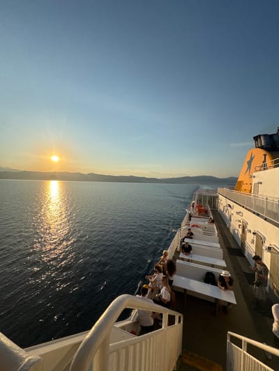 This image depicts a sunset over the Aegean Sea, viewed from the deck of a ferry in Kavala, Greece. The sun, a bright orb low on the horizon, casts a shimmering golden path across the dark blue water. The sky is a gradient of clear, light blue at the top, transitioning to a pale yellow and orange near the horizon, suggesting the warm glow of sunset.
The ferry's deck is visible, with white railings and seating areas. Several people are scattered across the deck, some sitting on benches and others standing, looking out at the sea or conversing. Their attire suggests a casual, relaxed atmosphere. The ferry itself appears to be a passenger vessel, with a white superstructure and some orange accents on the stern, including a large blue star logo.
In the distance, a silhouetted mountain range forms a backdrop to the scene. The water is calm, with gentle ripples reflecting the sunlight. The overall mood is serene and peaceful, capturing a moment of travel and contemplation as the day ends. The time of day is clearly sunset. There is no visible text in the image.