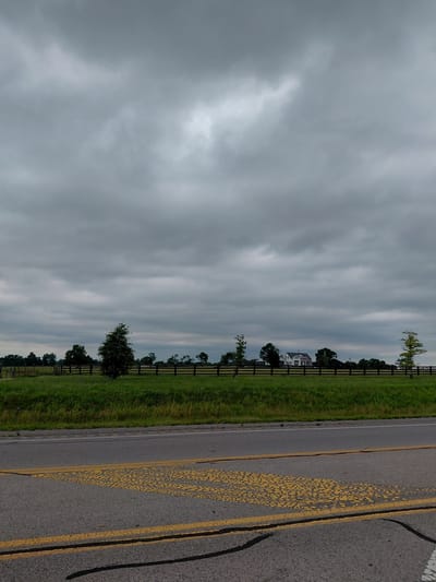 The image captures a rural roadside scene. The perspective is from a low angle, with the camera tilted sideways, making the sky and the ground appear vertical. The sky is overcast with thick, grey clouds, suggesting a gloomy or approaching storm.

To the left of the image, a vast expanse of sky dominates, filled with textured, layered clouds. In the distance, a white house with a dark roof is visible, nestled amongst trees and bordered by a dark wooden fence. This fence runs horizontally across the middle ground, separating a lush green grassy field from the road. Scattered trees are interspersed along the fence line.

On the right side of the image is an asphalt road. It features faded yellow double lines, with a section of broken, circular yellow markings. The road appears to be a two-lane highway. The overall impression is one of a quiet, overcast day in a rural setting, possibly in Danville, USA, given the provided location context. There is no visible text in the image.