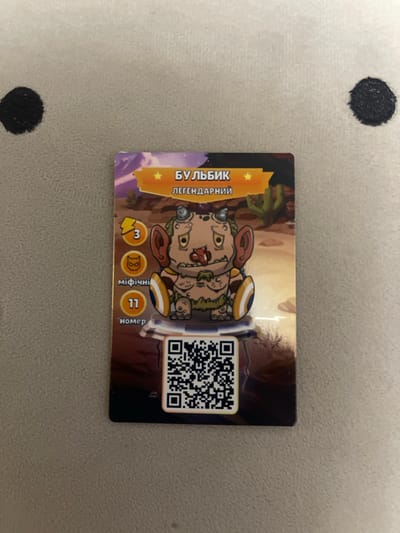 The image shows a collectible card with a cartoon character on it, laid flat on a textured surface. The card has a yellow banner at the top with Cyrillic text that reads "БУЛЬБИК ЛЕГЕНДАРНИЙ," which translates to "Bulbiyk Legendary." Below the text is an illustration of a creature with large ears, horns, and a yellow shield on its arm. To the left of the creature are three circular icons: a lightning bolt with the number 3, a cat-like mask, and a circle with the number 11. Below the number 11 are the Cyrillic words "міфічні" (mythical) and "номер" (number). At the bottom of the card is a QR code. The background of the card depicts a desert landscape with cacti. The textured surface underneath the card appears to be a grey fabric with black circular markings, resembling a playmat or a blanket. The image does not provide any information or visual cues that would allow for the identification of a specific location in Berezhany, Ukraine.