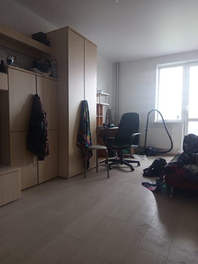 This is a medium-shot, eye-level photograph of a room. The room appears to be a living space or study, likely in an apartment in Kopeysk, Russia, given the overall aesthetic and the presence of common household items.
The scene is dominated by a light-colored wooden floor. To the left, there is a light-colored wooden closet or cabinet with two doors, one of which is slightly ajar. Hanging on this door is a dark jacket with a patterned scarf draped over it. Above this closet, a wooden shelf holds a few dark-colored boxes.
In the center of the room, a black office chair is positioned in front of a desk that is partially visible. The desk has some items on it, including what appears to be a pair of headphones and a book or magazine. To the right of the desk, a colorful, patterned fabric is draped over a thin metal chair with a small seat.
Further to the right, a large window with white frames dominates the wall, letting in a significant amount of light, which creates a strong highlight on the floor. Below the window, a white radiator is visible. A blue vacuum cleaner with a black hose is positioned on the floor in front of the radiator, with its hose extending towards the desk area.
In the far right of the image, a portion of a dark-colored sofa or chair is visible, covered with patterned fabric. A dark bag and some other fabric items are on the floor in front of this seating. The walls are painted a light, neutral color, and the ceiling is also light. The room appears to be somewhat cluttered but organized. The lighting suggests it is daytime, possibly late afternoon, given the strong directional light.