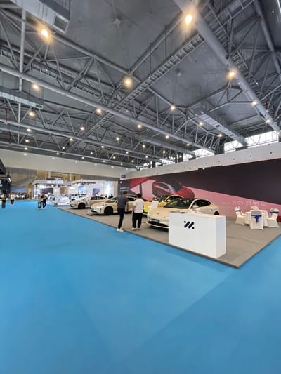 The image shows an indoor automotive exhibition with a blue carpeted floor and a large white display stand with the logo "W".  There are several cars parked on the floor and several people walking around.  The cars are all silver and white and are positioned in a line facing the white display stand. The background shows a large billboard displaying a silver car and the date "21.99-28.99". There are multiple white overhead lights in the large, industrial space with a metal ceiling. The location is the Hangzhou International Expo Center, based on the provided coordinates (30.272, 120.416).  There is a large white display stand with the logo "W" and several white chairs with blue fabric cushions. The cars are all on a blue carpeted floor.