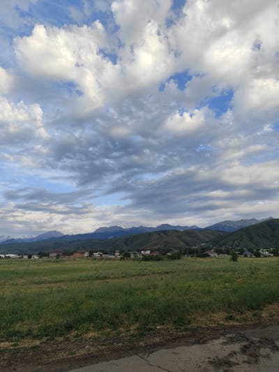 The image shows a landscape with a cloudy sky, green hills, a village, and a mountain range. The foreground features a dirt road and a grassy field. The village consists of several houses and buildings scattered across the foothills. The mountains in the background are partly covered in snow, indicating a high altitude. The sky is filled with large, white and gray clouds, with patches of blue sky visible. The scene appears to be in a rural or semi-rural area, possibly a suburb of Dolan, Kazakhstan, given the presence of a village and the characteristic mountain backdrop. There are no people or animals visible in the image. The lighting suggests it is either early morning or late afternoon.
