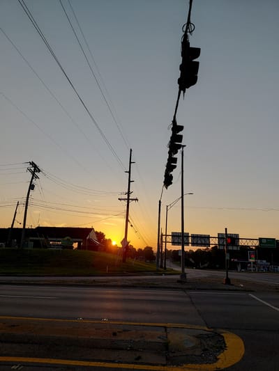 The image captures a sunrise over a commercial area in Frankfort, United States of America. The sky is a gradient of pale blue to soft orange, with the sun just beginning to peek over a grassy hill in the mid-ground. Several power poles with a complex network of wires crisscross the frame, silhouetted against the bright sky. In the foreground, a two-lane asphalt road with yellow dividing lines and a yellow painted curb is visible. On the right side of the road, a green highway sign indicates "460 EAST Georgetown." Beyond the hill, the tops of buildings and trees are visible, with a gas station and convenience store clearly identifiable on the right side. Traffic lights, also silhouetted, hang over the intersection. No people are visible in the scene.