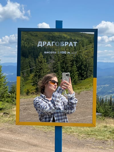 In the image, a young woman with brown hair and sunglasses is taking a selfie with her smartphone. She is wearing a blue and white plaid shirt and standing in front of a reflective sign. The sign displays the text "ДРАГОБРАТ" and "висота 1400 м" in white lettering. Behind her, a mountainous landscape with lush green trees is visible. The sky is blue with scattered white clouds. The sign is mounted on a blue and yellow pole, with a yellow frame around the reflective surface. The reflection shows the woman holding her phone and the surrounding environment. The location is identified as Dragobrat, in the city of Chorna Tysa, Ukraine.