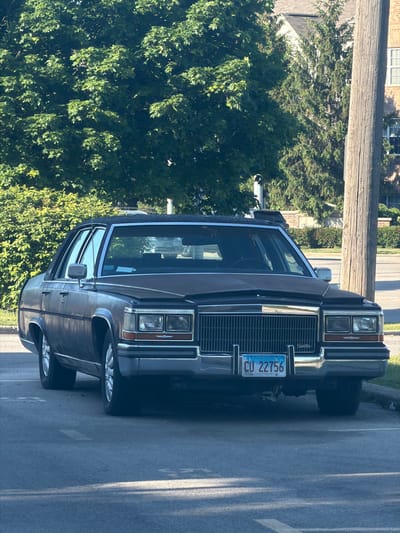 The image shows a dark-colored Cadillac Brougham parked on a paved road or street in Morton Grove, Illinois. The car is facing the viewer. The Illinois license plate "CU 22756" is visible on the front of the car. A wooden utility pole is situated nearby. Green foliage surrounds the car, and there appears to be a building in the background.