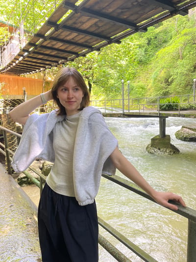 A young woman is captured in a daylight outdoor setting in Tsabal, Abkhazia. She stands on what appears to be a walkway or bridge, leaning her right arm on a sturdy metal railing that overlooks a vibrant, fast-flowing river. Her left hand is raised towards her head, subtly touching her hair or neck. She has light brown hair, a calm, pleasant expression, and is looking directly at the camera with a slight smile.
She is dressed in casual attire, wearing a light grey oversized hooded sweatshirt and a dark, possibly black, flowing skirt. A light-colored wristwatch is visible on her left wrist.
The background behind her features a dynamic natural landscape. To her right, the river, appearing light green or slightly murky, rushes past with visible rapids and scattered rocks. Beyond the riverbanks, dense, lush green foliage and trees dominate the mid-ground, indicating a rich, verdant environment. To her left and spanning behind her, a rustic wooden bridge with dark wooden planks, metal supports, and rope railings is visible, extending over a leafy canopy. Another wooden structure or building can be glimpsed further back on the left side. The lighting is bright and natural, suggesting a clear or lightly overcast day. There is no visible text in the image.