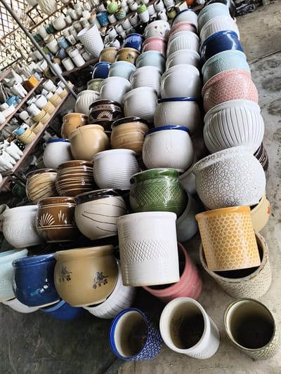 The image shows a large collection of ceramic plant pots stacked on the floor and displayed on shelves, likely for sale. The pots vary in size, shape, color, and design. The scene appears to be in a shop or warehouse environment, with shelves filled with even more pots visible in the background. The location is Ghaziabad, India. No people are visible.