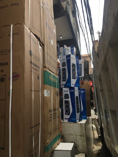 The scene depicts a cluttered, narrow exterior passage between buildings in Ojo, Nigeria, consistent with a commercial or storage area. The foreground and midground are dominated by stacks of packaged merchandise. On the left, several large, brown cardboard boxes are stacked vertically, secured with white strapping. Visible branding includes "GLAMSTAR" for a "REFRIGERATOR" (model FR-175DT-W) and "Hisense" for a "Washing Machine." One box indicates a weight of "23 kg." On the right, a rough, light-colored concrete wall forms one side of the passage. Adjacent to it, blue and white branded boxes for "ALIPU" speakers are stacked on a low, white concrete ledge. These speaker boxes display images of black speakers with prominent circular, multi-colored light rings. A white plastic bag rests on the topmost ALIPU box. Further back on the right, additional appliance boxes from brands like "EXLG" and "SONOS" are visible, with a glimpse of a person in dark clothing near a red sign. The upper background reveals a bright sky above the gap between buildings, traversed by numerous dark, loosely hanging electrical wires. A small light fixture is visible on a building facade in the upper center. No overt actions or interactions are discernible among people.