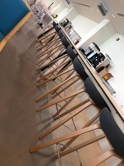 The image depicts an interior view of a modern, long, and narrow commercial space located in Hanak, Saudi Arabia. The primary focus is a lengthy, light-colored wooden bar or counter extending diagonally from the mid-right foreground into the background. A row of approximately eleven dark grey upholstered bar stools with light wooden frames and chrome footrests is aligned along the counter.
Behind the counter, various commercial beverage preparation equipment is visible, including multiple espresso machines or coffee makers, a grinder, and blenders. A metal shelving unit and an open cardboard box are also present. The floor is a smooth, light brown or greyish-brown material. The walls are white, and the ceiling is equipped with recessed lighting and two overhead air conditioning or ventilation units.
In the background, on the far left, a section of a blue upholstered bench or seating area is partially visible. One individual is present in the mid-background, standing behind the counter. This person is wearing a neon yellow or green vest over dark clothing and appears to be involved in work or setup, as suggested by the presence of a ladder and other items in the background that suggest an ongoing installation or preparation phase. The overall scene indicates a contemporary establishment, possibly a cafe or breakroom, in a state of completion or readiness.