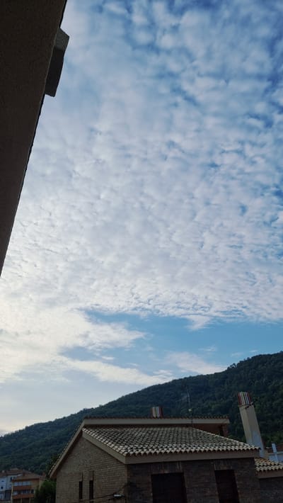 The image captures a view from a building in Sant Joan Les Fonts, Spain, looking upwards towards a cloudy sky and a tree-covered mountain. In the foreground, a portion of a building's exterior wall, made of light-colored material, is visible on the left side. Below this, a roof with terracotta tiles covers a building constructed of brick. This building features dark rectangular openings, possibly windows or doorways, and a chimney with colorful vertical stripes. To the right, another chimney with similar colorful stripes extends upwards. The background is dominated by a steep, lush green mountainside, densely covered with trees. The sky is a light blue, filled with scattered, fluffy white clouds. In the distance, some buildings with light-colored facades and orange roofs can be seen at the base of the mountain. An antenna is visible on the mountainside above the building with the tiled roof.