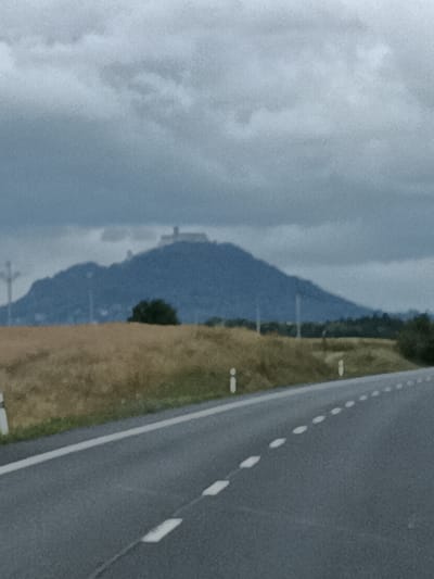 The image depicts a landscape scene with a road in the foreground, a field of dry grass in the middle ground, and a large hill with a structure on top in the background. The sky is overcast with clouds. The road has white dashed lines indicating lanes. The hill appears to be a prominent natural feature with a man-made structure, possibly a castle or observation point, situated on its summit. The vegetation in the middle ground is predominantly dry and appears to be a field of crops or grassland. There are a few scattered trees and some utility poles visible. The overall impression is one of a rural or semi-rural setting under a cloudy sky. The presence of a road suggests it is likely a view from a vehicle traveling along a highway or main road.
